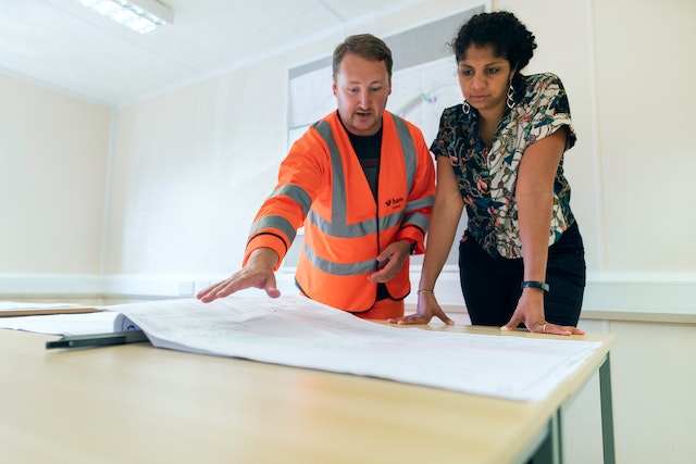 a contractor in an orange reflective vest looking over blue prints with an owner